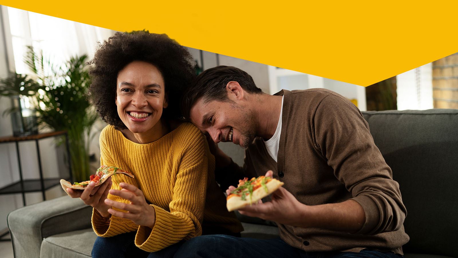 Two people eating pizza together on the sofa (with the yellow design element overhanging)