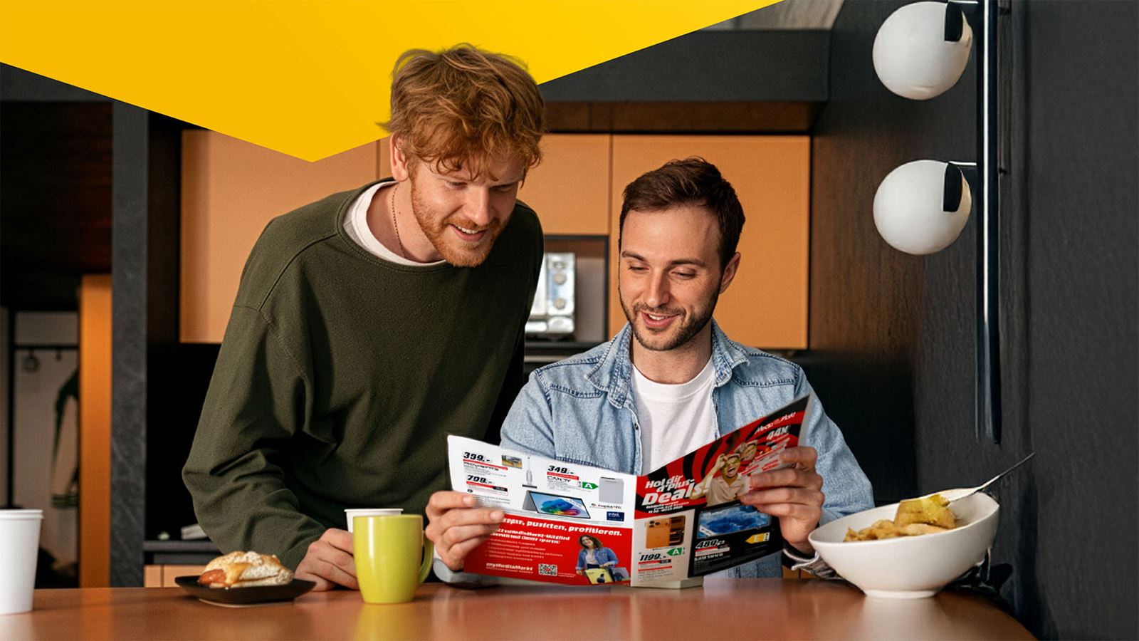 Deux jeunes hommes étudient un catalogue de produits de MediaMarkt à la table du petit-déjeuner (avec un élément de design jaune au-dessus)