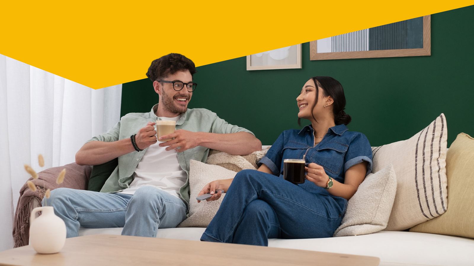 Un homme et une femme prennent un café sur un canapé (avec élément de design jaune au-dessus)
