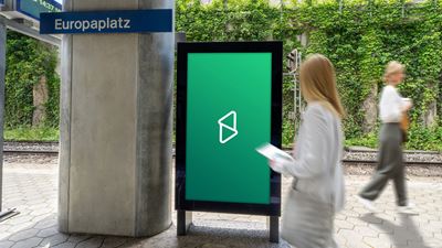 Schermo pubblicitario digitale in una stazione, con alcune passanti in movimento in primo piano; sullo sfondo si vedono i binari e un muro ricoperto di vegetazione.