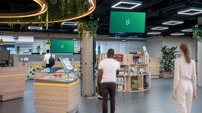 Vista interna di un moderno spazio di vendita o di permanenza con diversi schermi digitali sospesi al soffitto; in primo piano si vedono persone in movimento e varie superfici espositive.