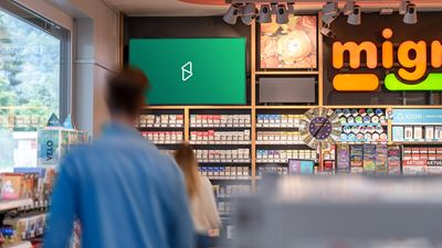 Schermo pubblicitario digitale nell’area vendita di un chiosco o di un convenience shop; in primo piano si vedono clienti sfocati e scaffali con prodotti.