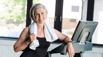 An elderly, sporty woman with sportswear and a towel stands in front of a home trainer in a gym.
