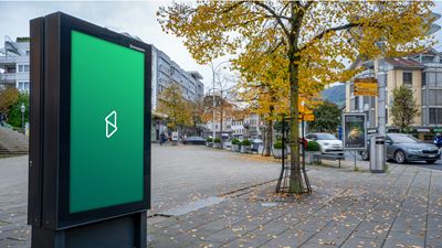 Schermo pubblicitario digitale nello spazio pubblico, lungo una strada o in un centro urbano; sullo sfondo si vedono alberi, edifici e auto di passaggio.