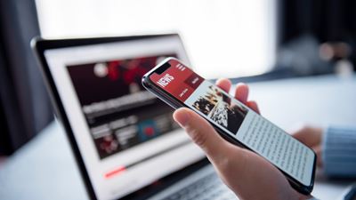 Jemand hält ein Smartphone, im Hintergrund ist ein Bildschirm